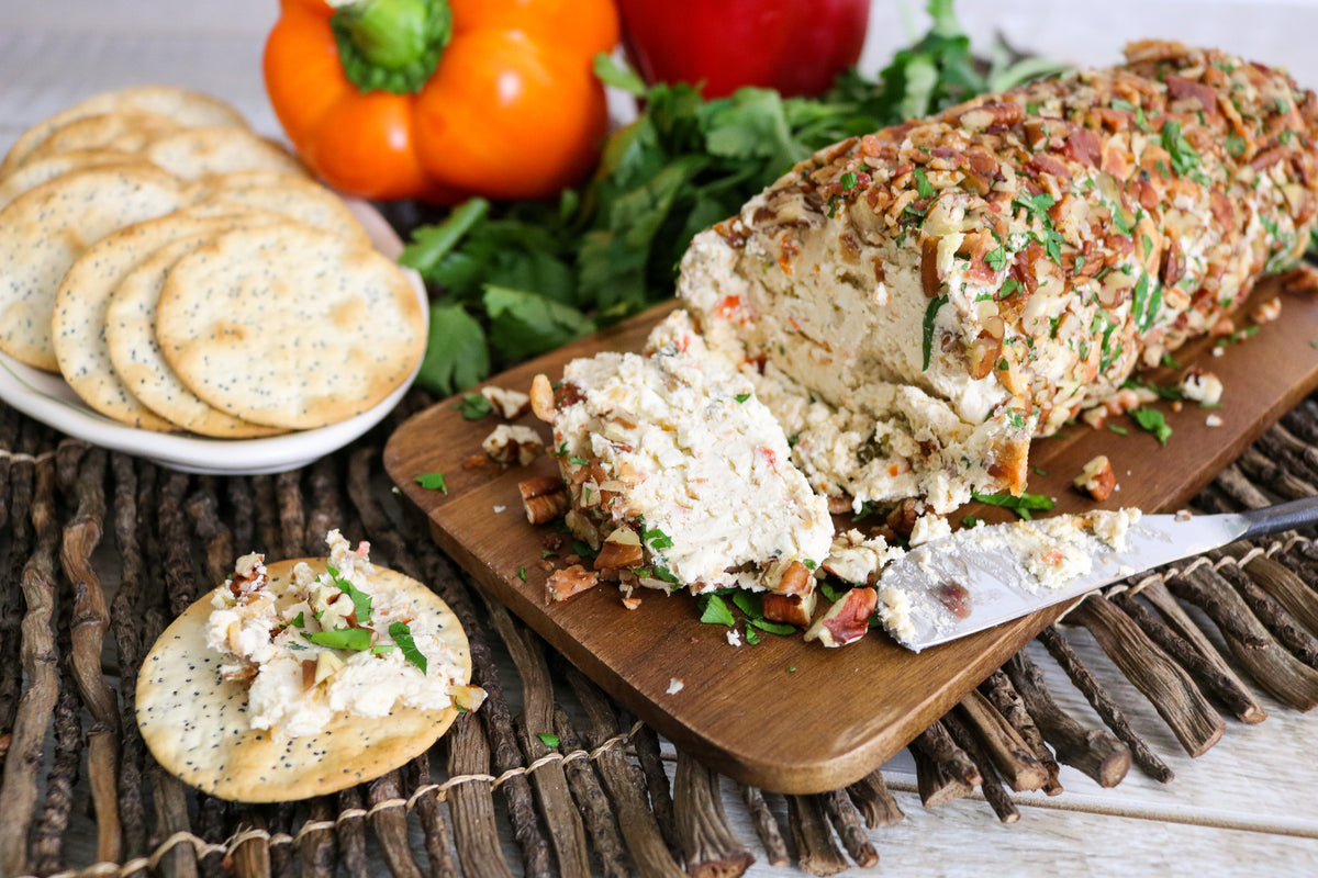 A sliced cheese and bacon stuffed log cake on a wooden cutting board, served with crackers on a rustic table setting. Fresh parsley, red bell peppers, and a white plate of round crackers are visible in the background.