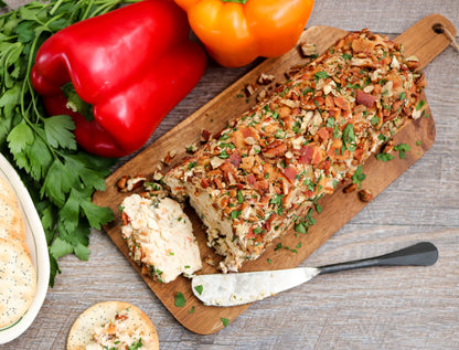 cheese on a wooden cutting board, garnished with chopped herbs and crispy bacon bits, surrounded by fresh red and orange bell peppers, parsley, and a knife on a gray wood surface. garden party mix