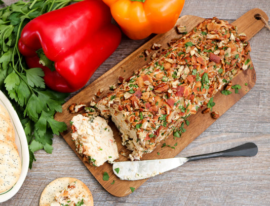 cheese on a wooden cutting board, garnished with chopped herbs and crispy bacon bits, surrounded by fresh red and orange bell peppers, parsley, and a knife on a gray wood surface. garden party mix