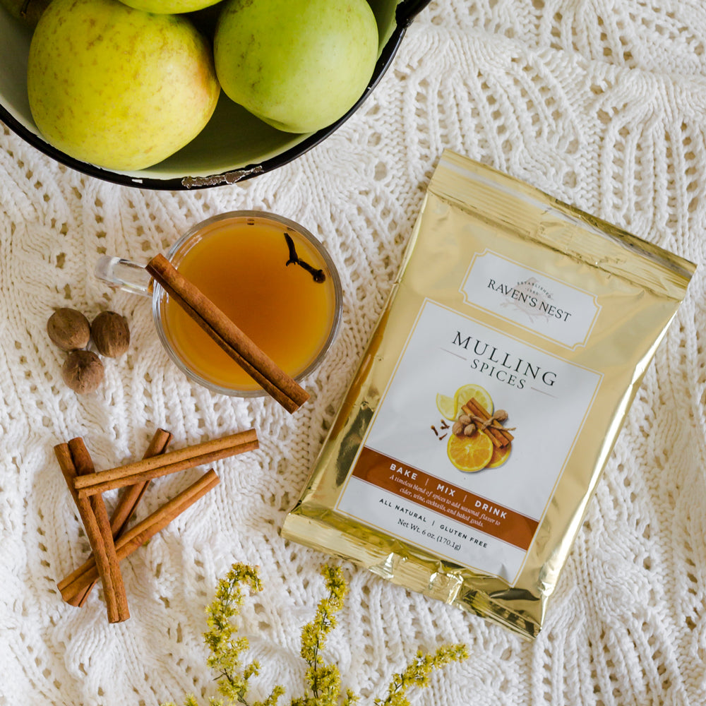 Mulling spice package by Raven's Nest on a crochet cream tablecloth with mulled cider, apples, and spices