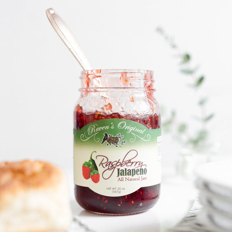 Open jar of Raven's Original raspberry jalapeno jam with a stack of plates and biscuits and a spoon