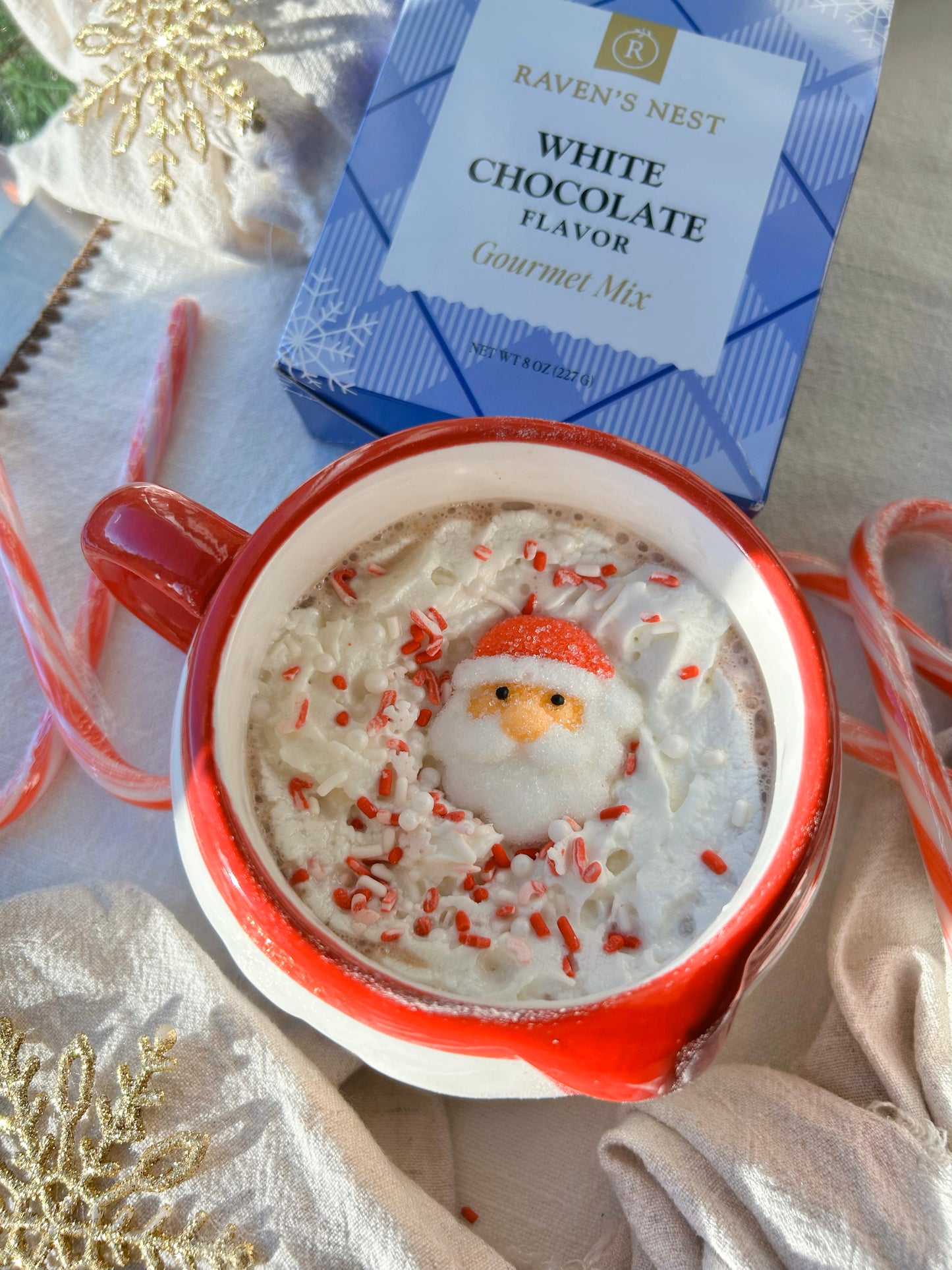 White Hot Chocolate garnished with whipped cream, holiday sprinkles and Santa candy in a festive mug surrounded by candy canes and snowflake ornaments