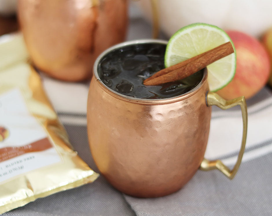 Copper cup with spiced apple cider moscow mule garnished with cinnamon stick and lime and package of Raven's Nest Mulling Spice